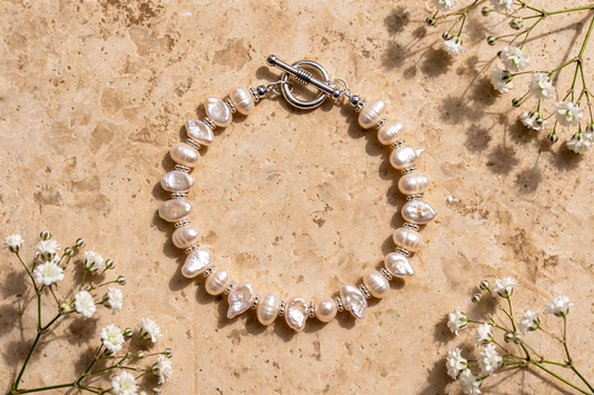 A beaded bracelet made of irregular white pearls separated by small silver spacer beads, with a silver toggle clasp, arranged in a circle on a beige stone surface with small white flowers around it.
