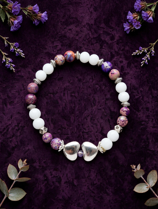 A beaded bracelet arranged in a circle on a dark purple textured background, featuring white beads, purple marbled beads, silver spacer beads, and two silver heart charms at the front.