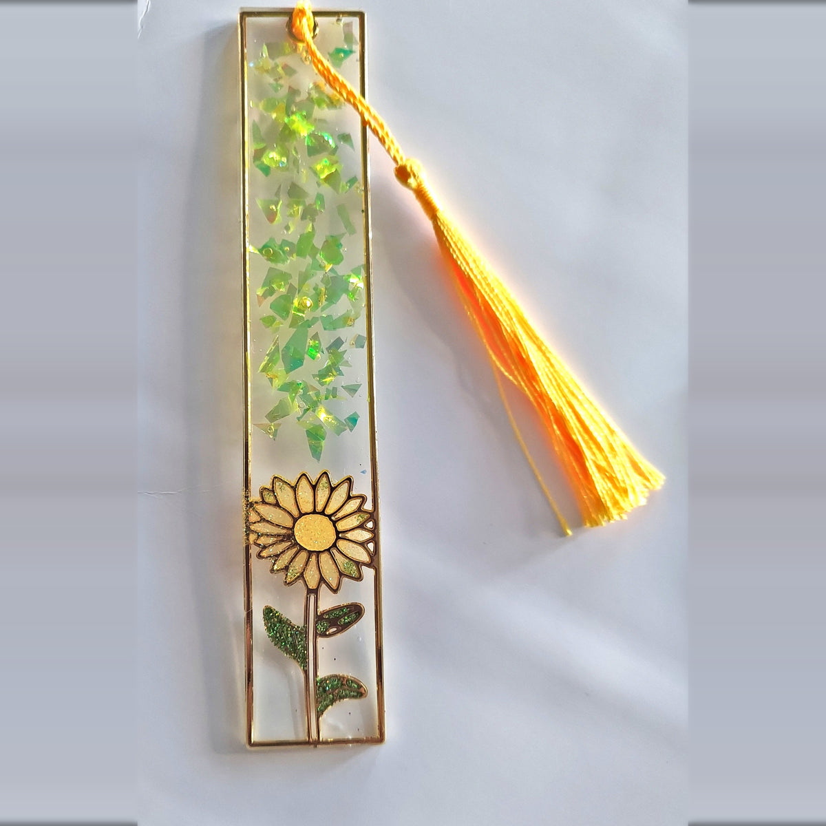 Handmade yellow resin bookmark with sunflower design, iridescent glitter flakes and yellow tassel on a light background
